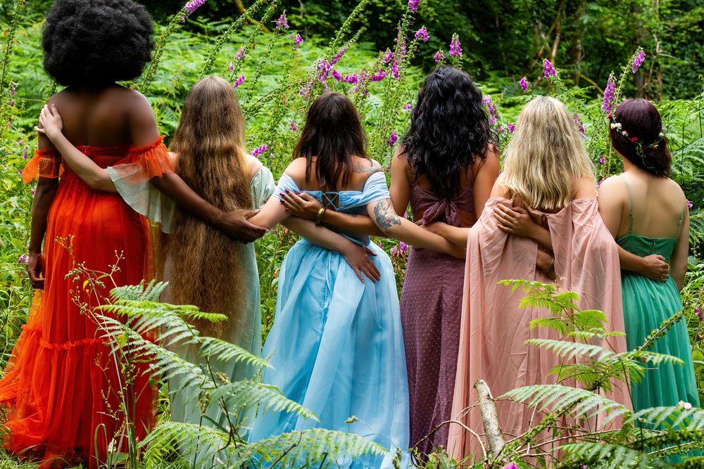 Six women standing shoulder to shoulder with their backs to the camera, arms around each other, among foxgloves