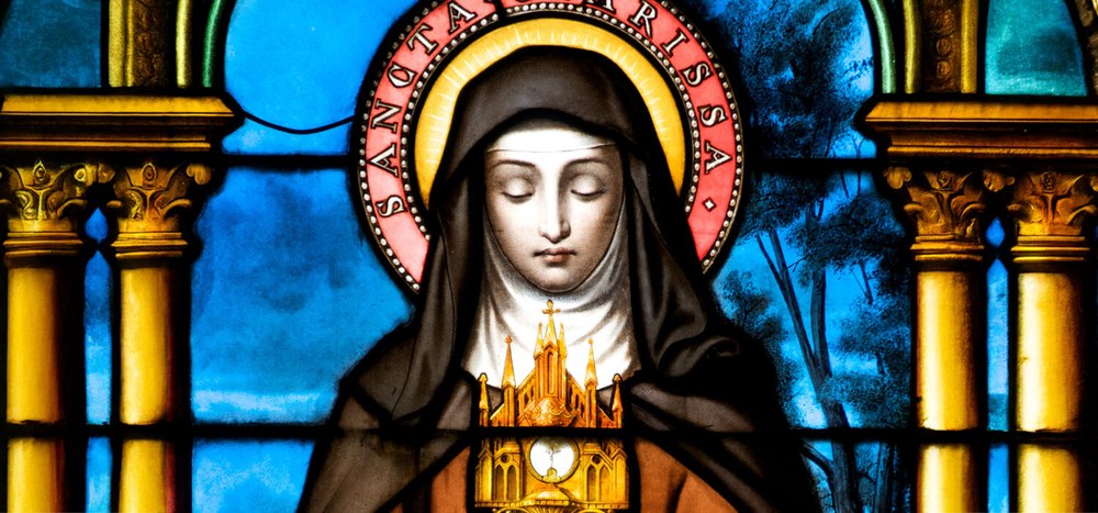 Clare of Assisi in stained glass: eyes closed, head bowed, her name written around the red halo