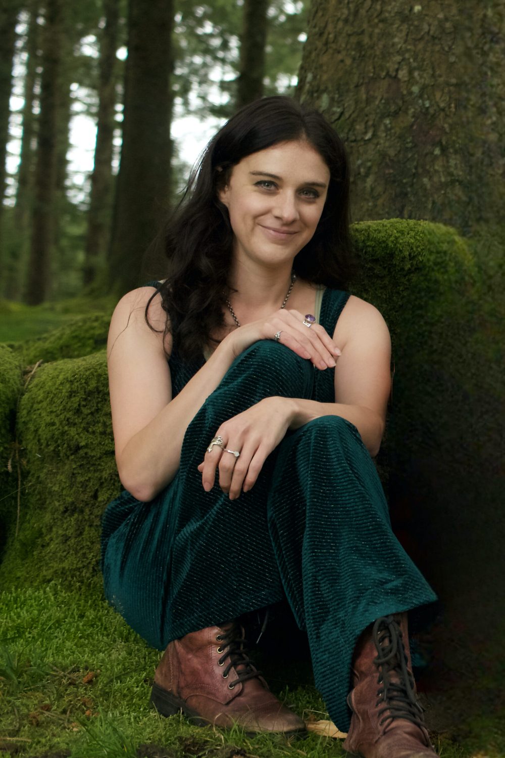 Ellie sitting against a mossy tree in a forest, boots planted, looking straight at the camera