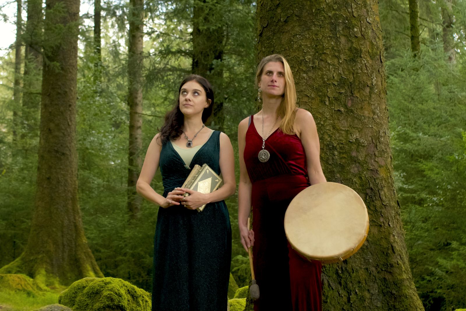Two women standing among the trees, one holding a frame drum, both looking up